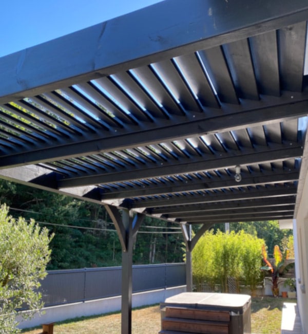 Pergola bioclimatique noire : lames ajustables et spa Pergola bioclimatique noire adossée à la maison, dotée de lattes ajustables, couvrant un spa sur la terrasse arrière ensoleillée.