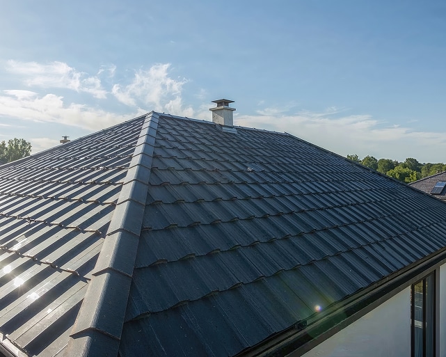 Toiture en tuiles métalliques anthracite pour maison moderne. Toit pyramidal en tuiles métalliques anthracite, très brillant. Ligne de croupe centrale et petite cheminée sous un ciel bleu dégagé.