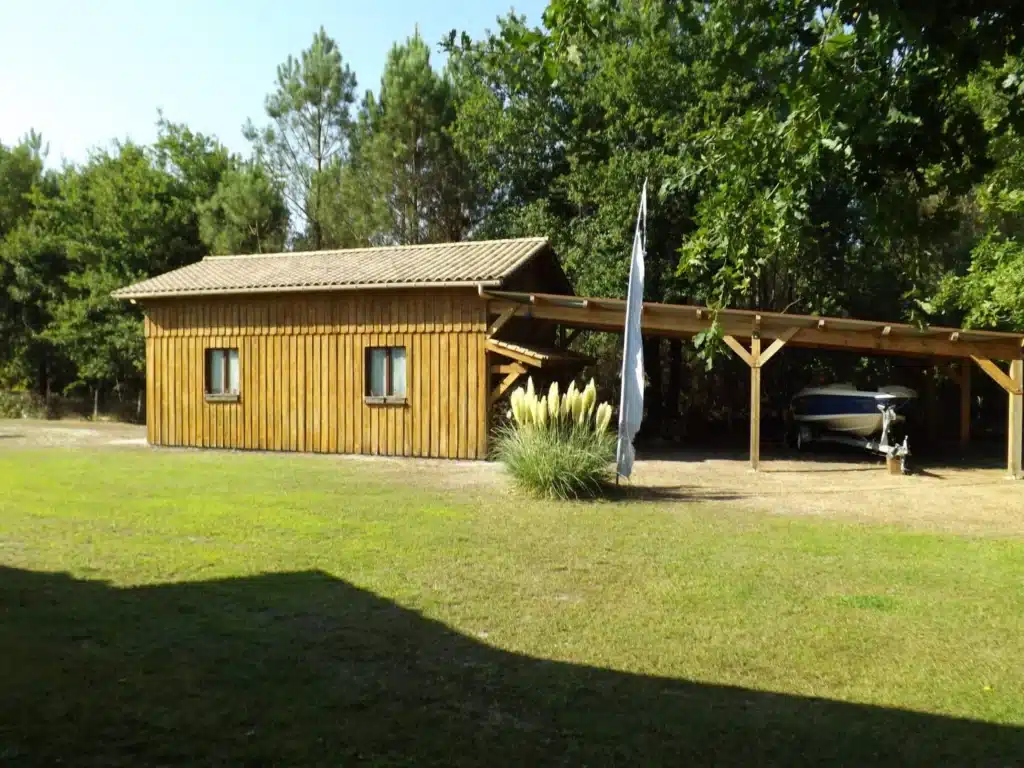 Chalet de Jardin en Bois avec Carport et Bateau Cabanon de jardin en bois et carport avec bateau sur remorque devant une forêt ensoleillée.