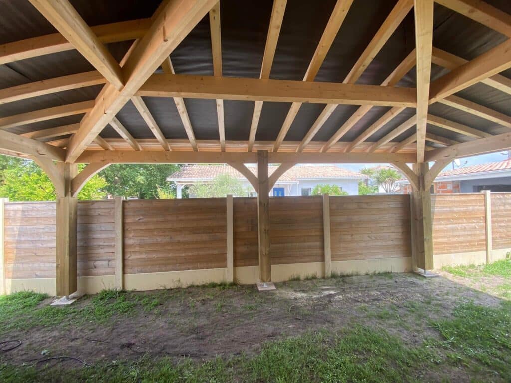 Charpente traditionnelle en bois massif pour une pergola de jardin avec poteaux robustes devant une clôture en bois.