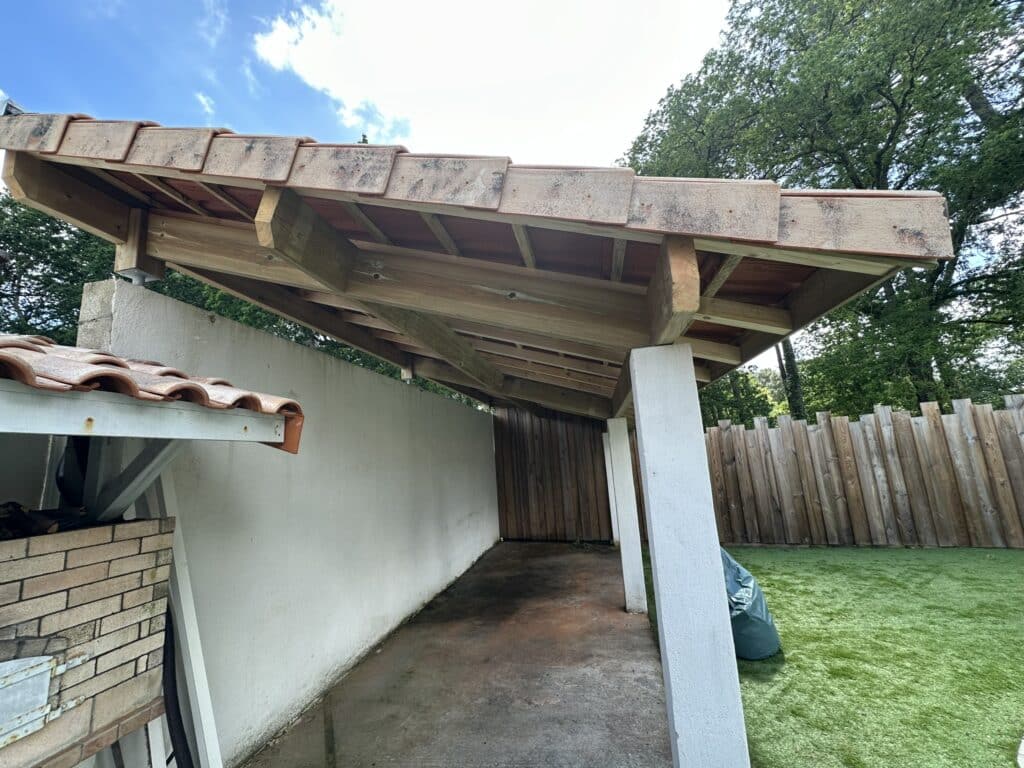 Abri de jardin avec toit en tuiles, poutres en bois apparentes, piliers blancs et pelouse synthétique.