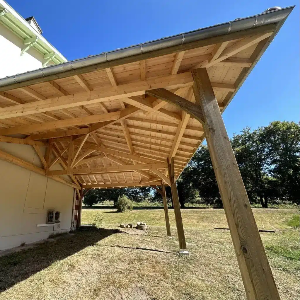Auvent en bois avec charpente traditionnelle sur mesure Auvent en bois massif avec charpente traditionnelle complexe, piliers inclinés et toiture tuilée devant un grand jardin.