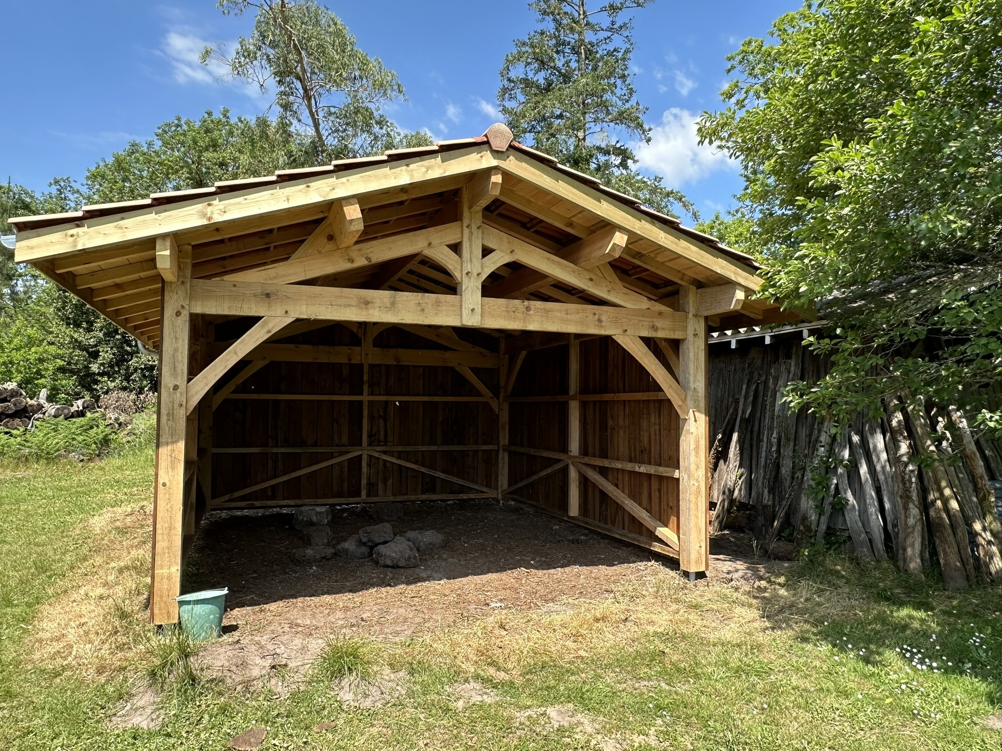 Abri de jardin ouvert en bois massif avec charpente traditionnelle et toit en tuiles sous un ciel bleu.