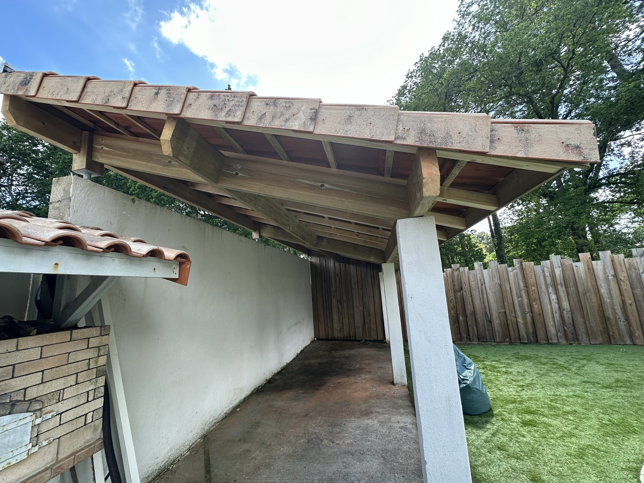 Abri de jardin avec charpente en bois robuste, toiture en tuiles et piliers blancs sur une pelouse synthétique.