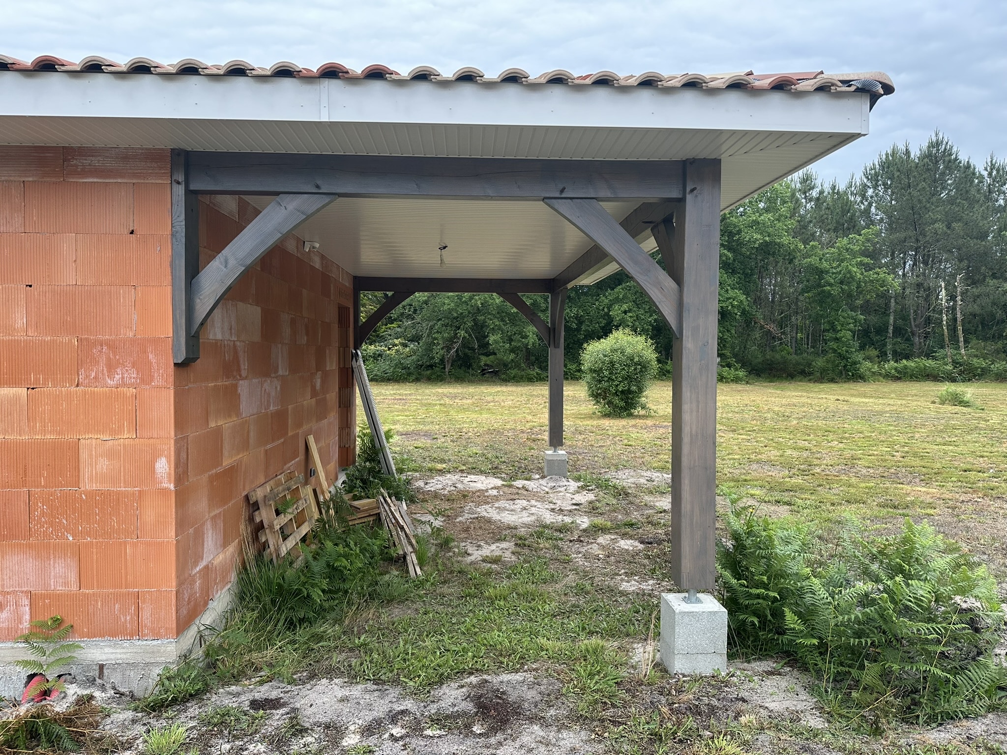 Maison en briques avec abri de terrasse en bois gris et toit en tuiles rouges dans un jardin.
