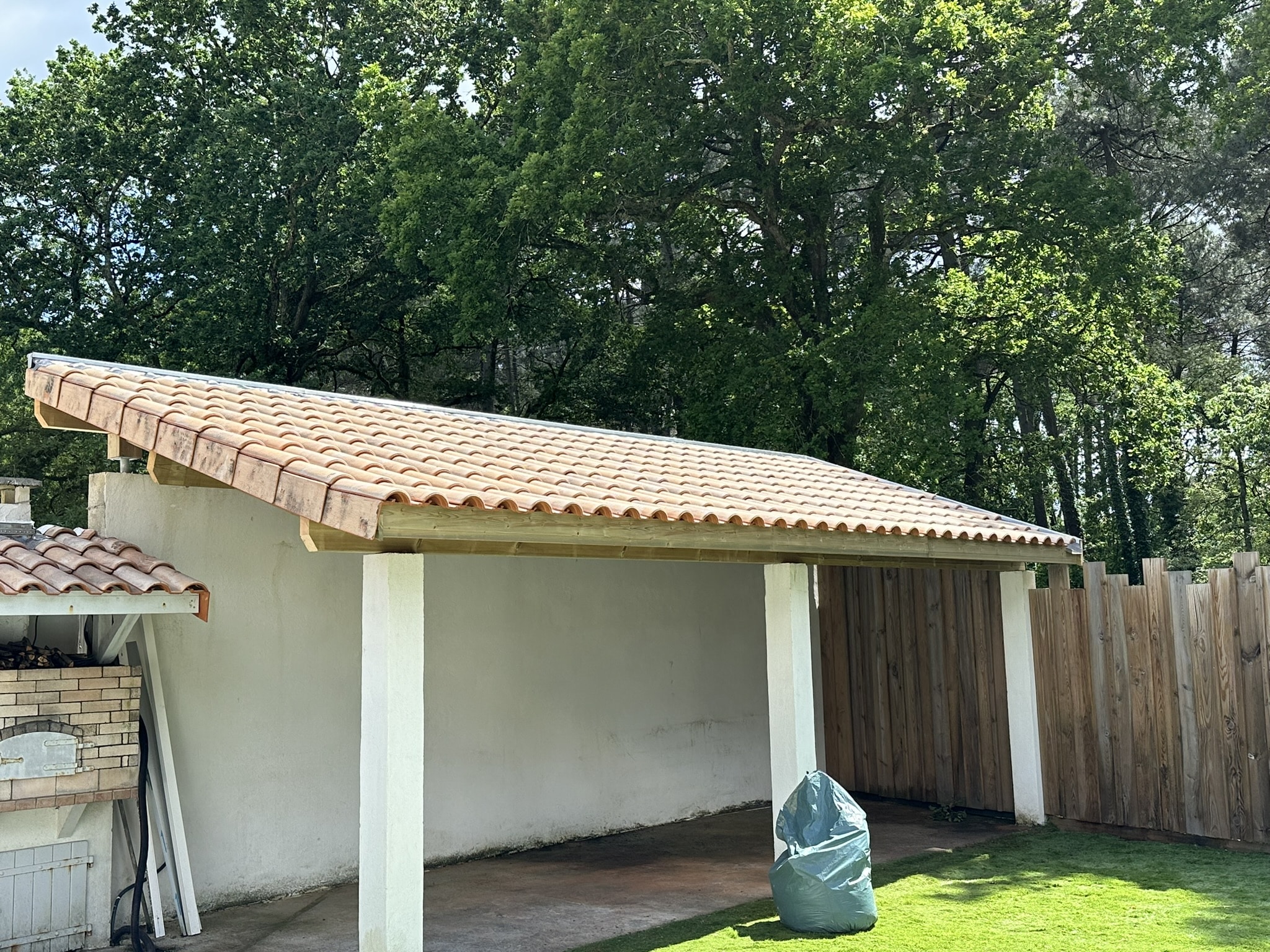 Abri extérieur avec toiture en tuiles romanes, murs blancs et structure en bois sur fond de forêt verdoyante.