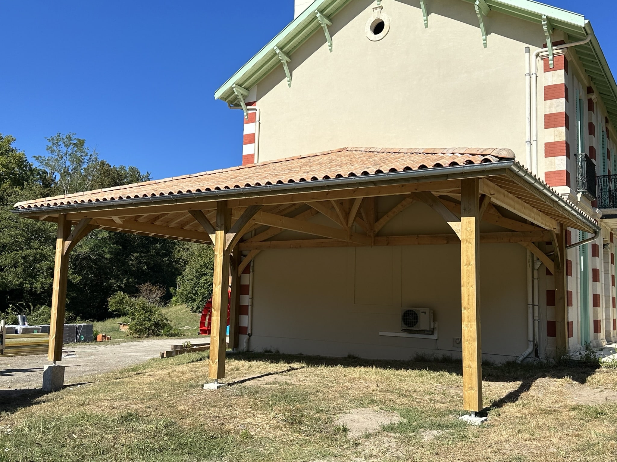 Carport en bois avec toiture en tuiles et charpente traditionnelle, attenant à une maison de caractère.
