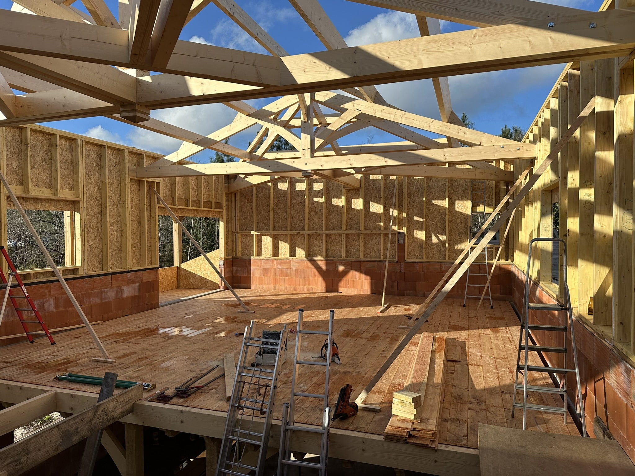 Chantier de construction avec charpente en bois apparente, murs en OSB et briques, et échelles sur le plancher.