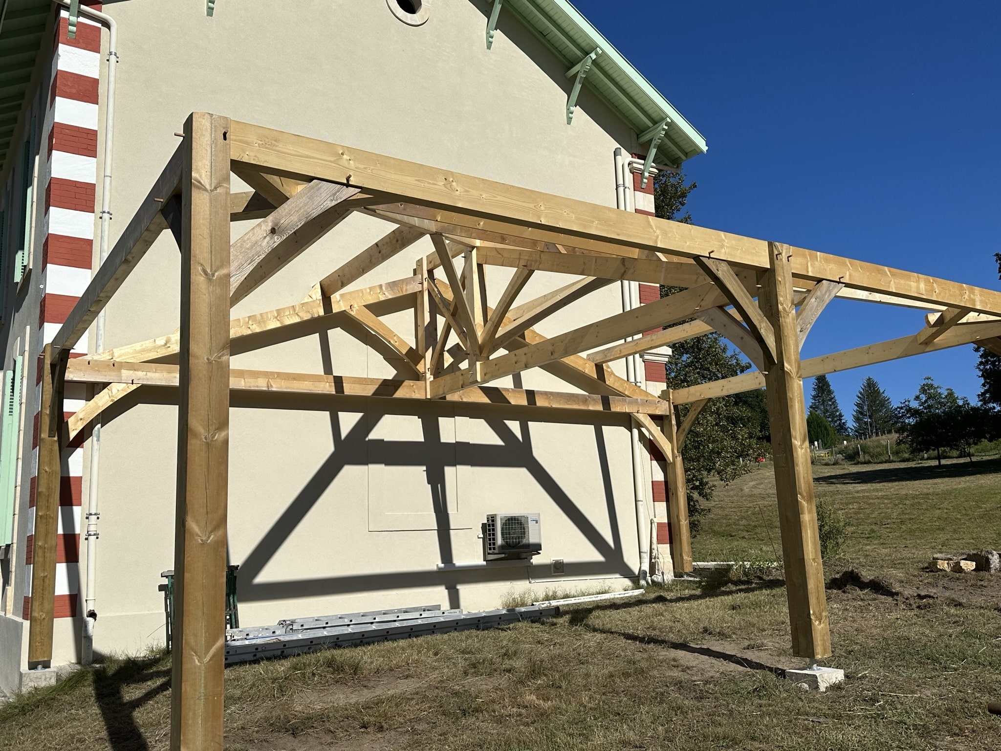 Charpente d'abri de voiture ou pergola en bois massif adossée à une maison sur un terrain en pente.