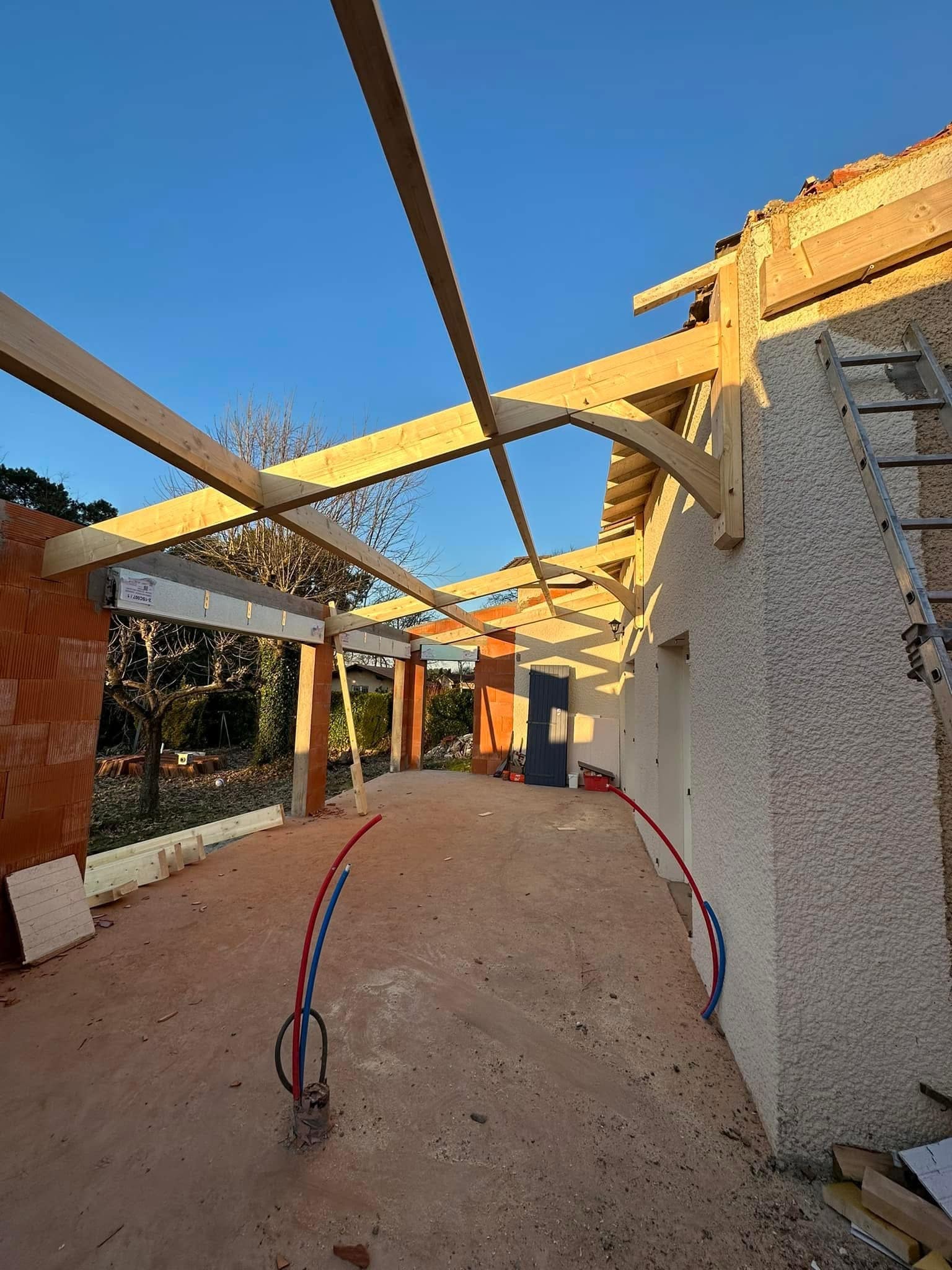 Charpente bois d'une extension de maison en construction avec murs en briques et tuyaux PER au sol.