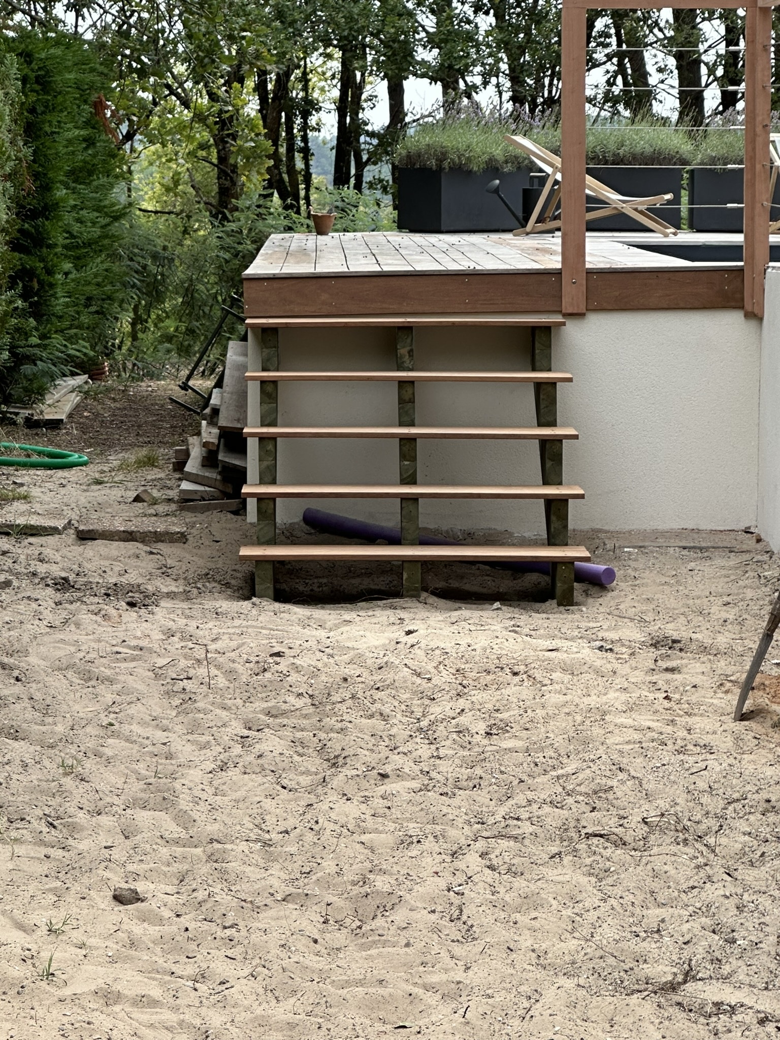 Escalier en bois clair en construction menant à une terrasse surélevée dans un jardin sablonneux.