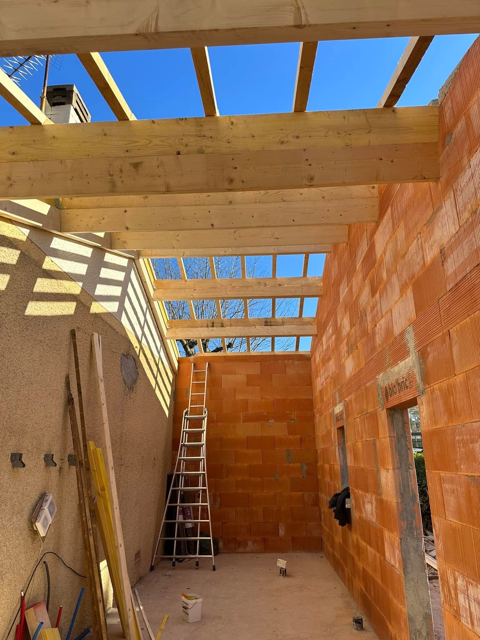 Charpente en bois apparente et murs en briques rouges d'une extension de maison sous un ciel bleu ensoleillé.