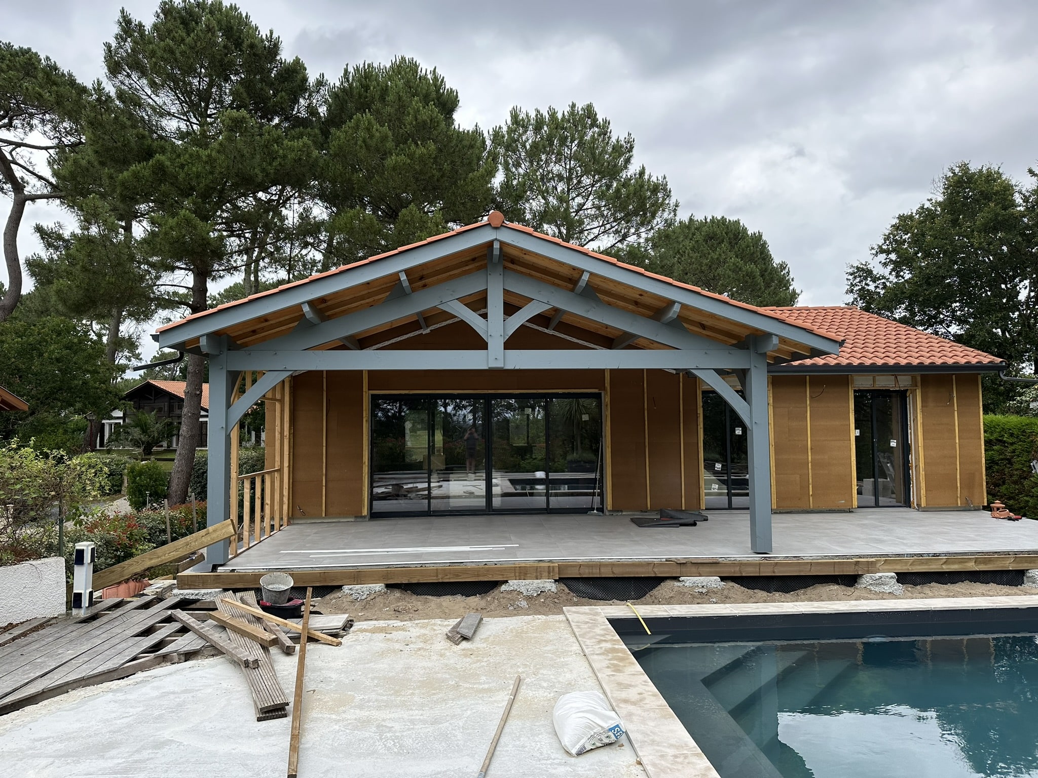 Maison en construction avec charpente bois grise, toit en tuiles et piscine moderne en premier plan.