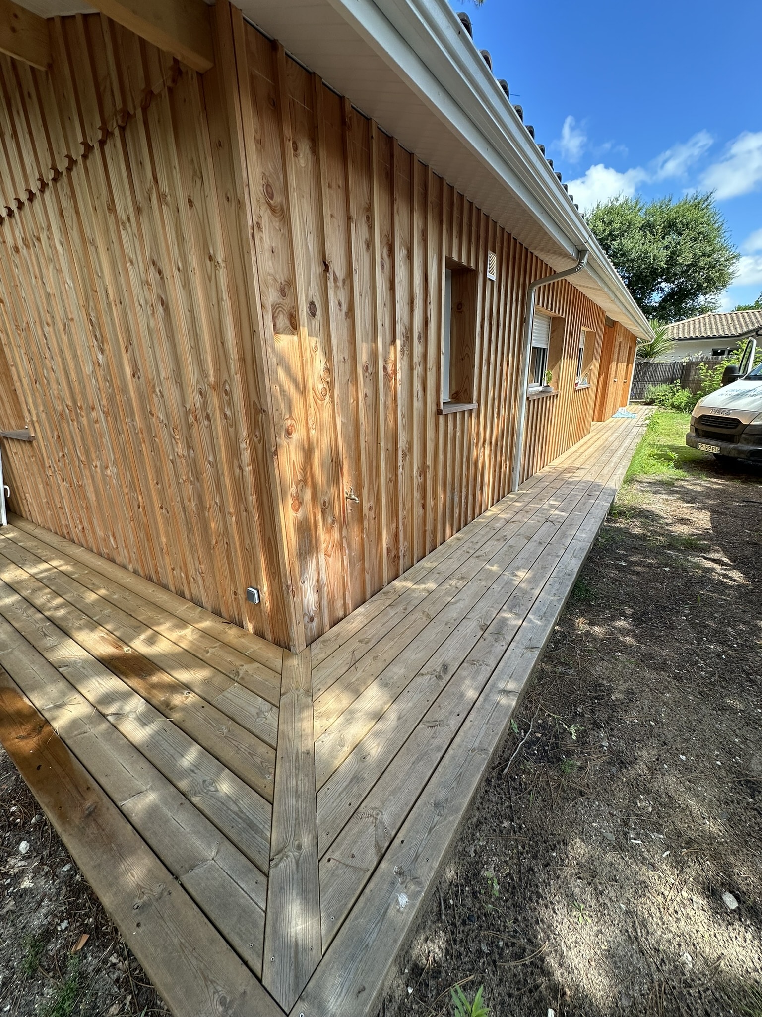 Maison avec bardage bois vertical et terrasse en bois massif, extérieur en bois naturel sous ciel bleu.