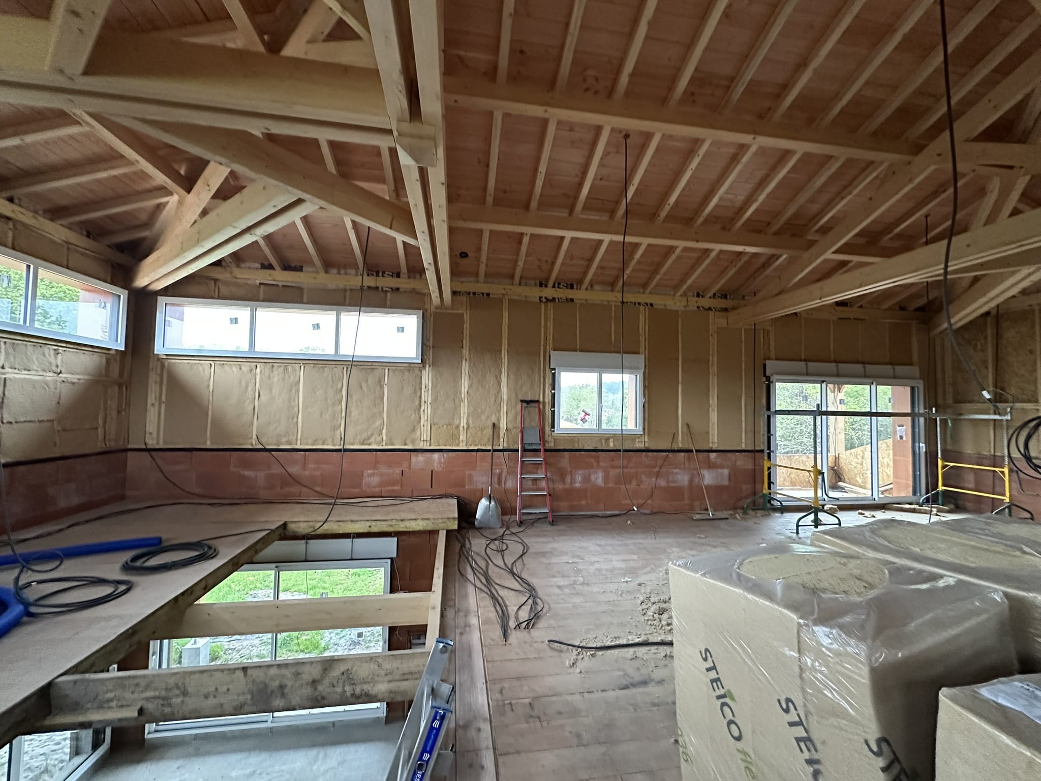 Chantier intérieur maison bois avec charpente apparente, isolation STEICO flex, fenêtres et briques.