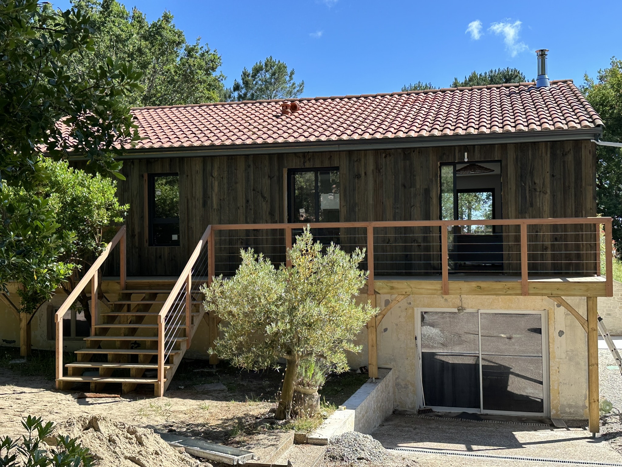 Maison en bois moderne avec terrasse surélevée, toit en tuiles rouges et escalier extérieur dans un jardin ensoleillé.