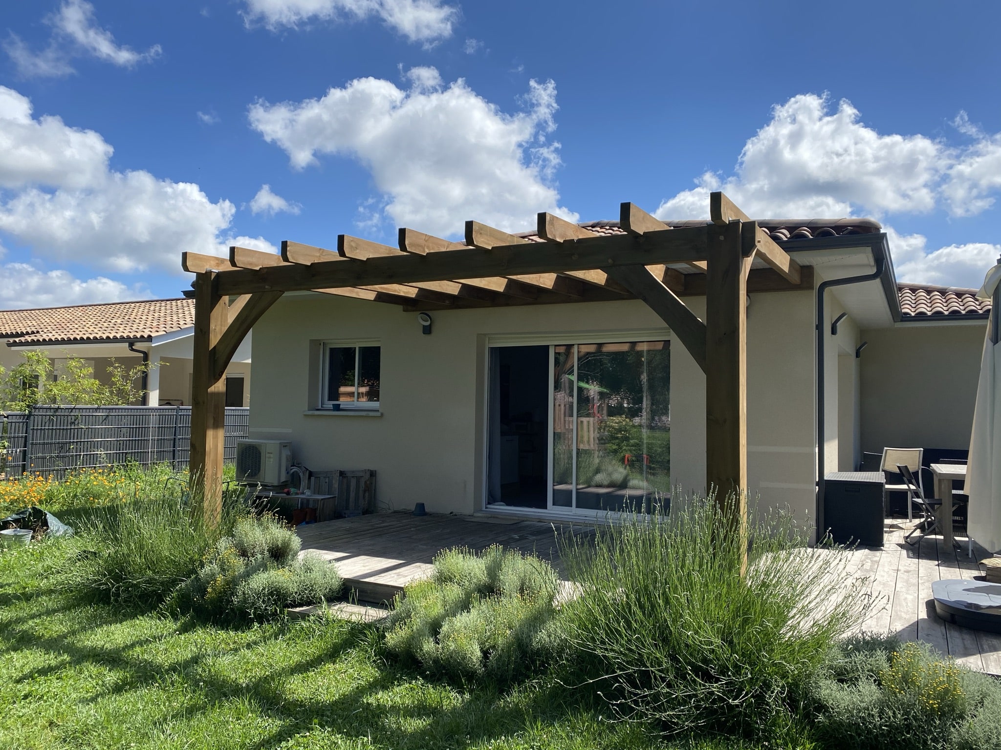 Pergola en bois sur une terrasse en bois devant une maison moderne, jardin ensoleillé sous un ciel bleu nuageux.