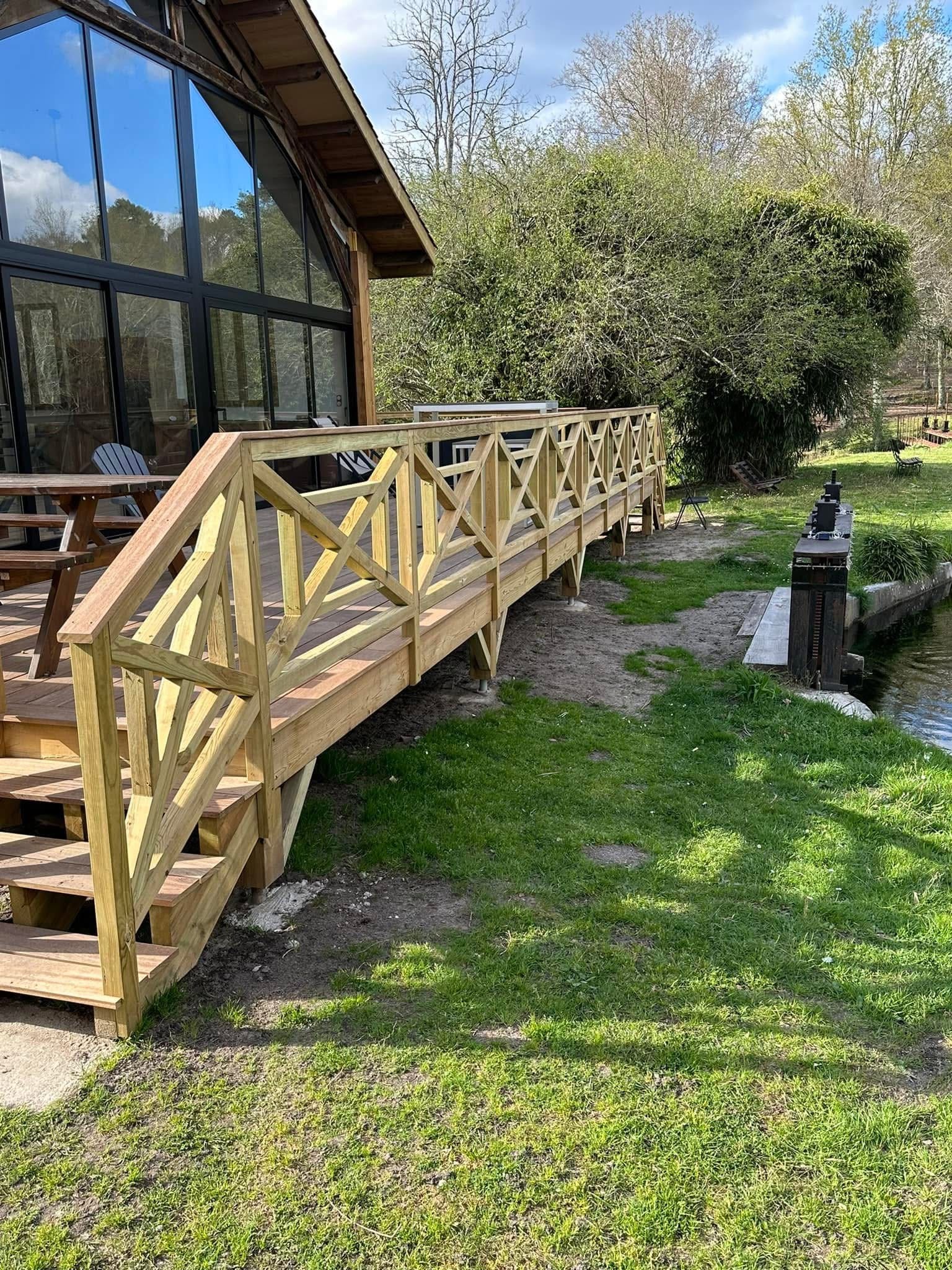 Terrasse en bois clair avec garde-corps en X bordant une maison vitrée moderne au bord d'un canal.