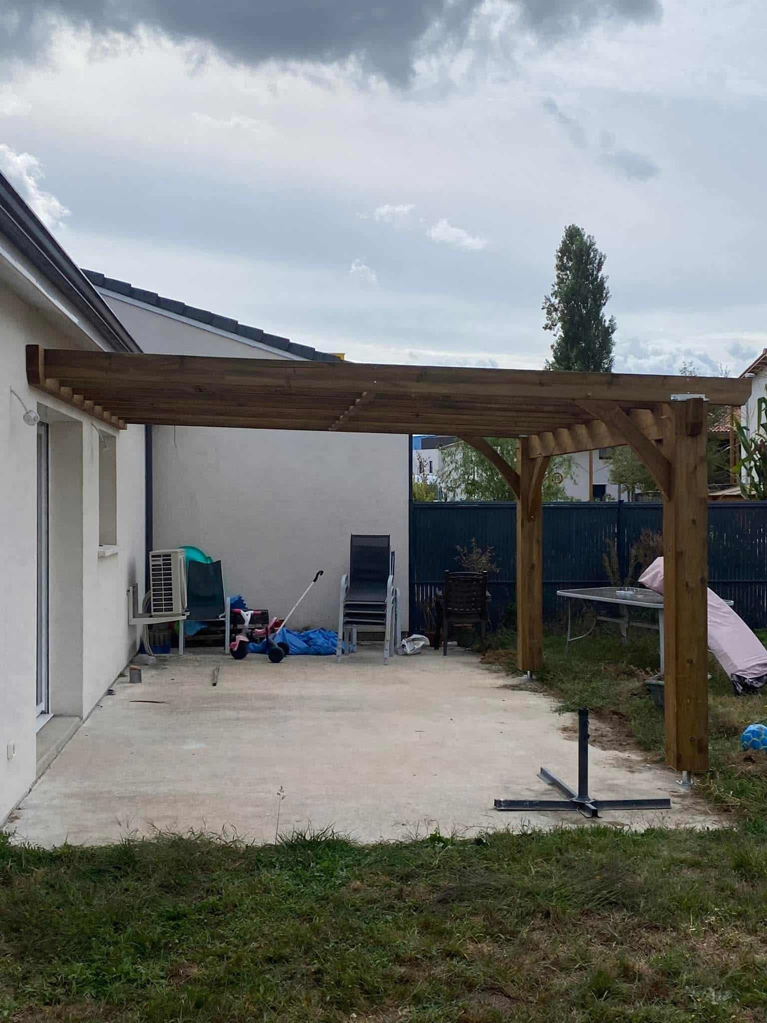 Pergola bois adossée à une maison sur terrasse béton avec mobilier de jardin.