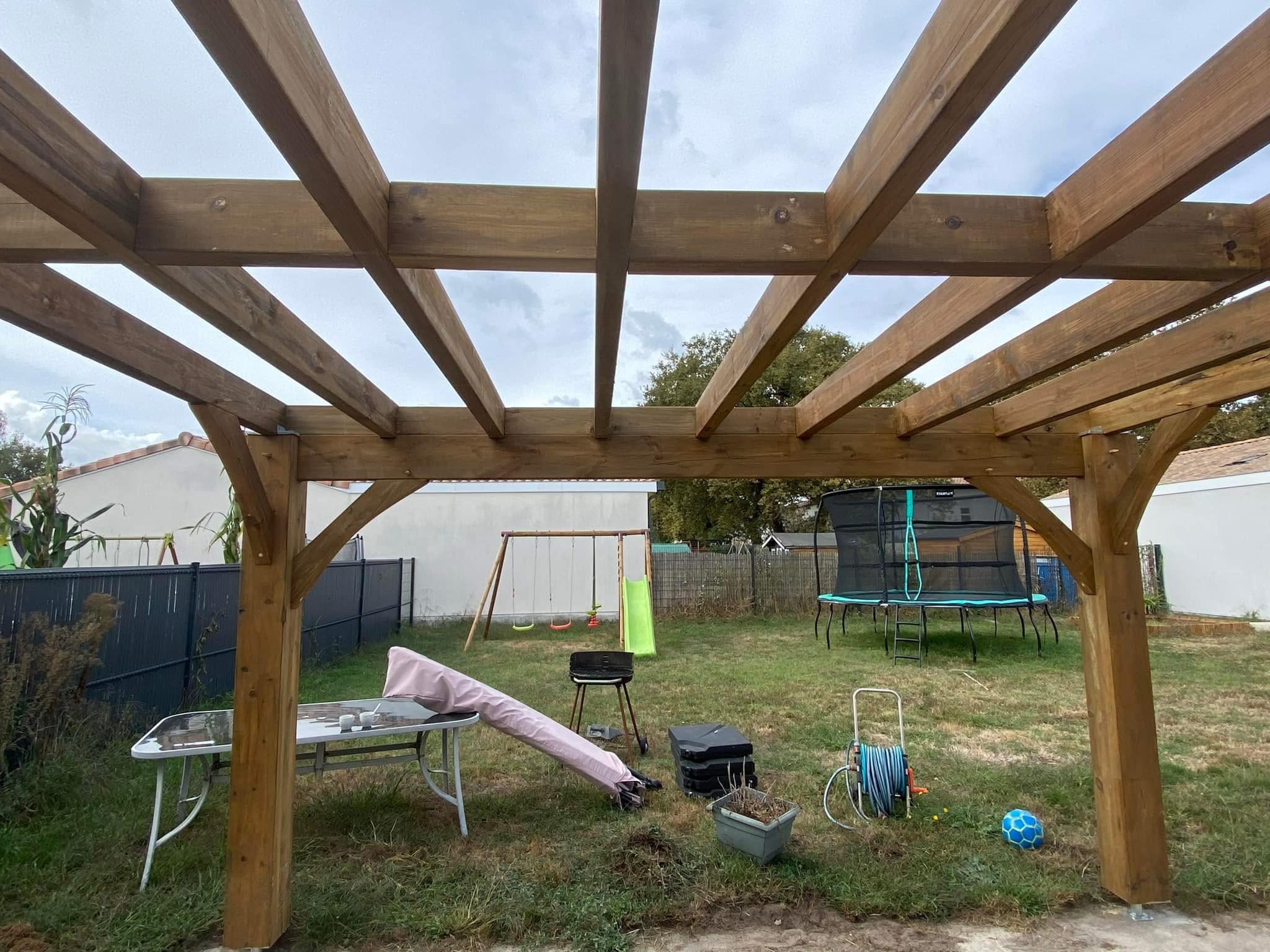 Pergola bois robuste surmontant un jardin avec trampoline, balançoire et barbecue.