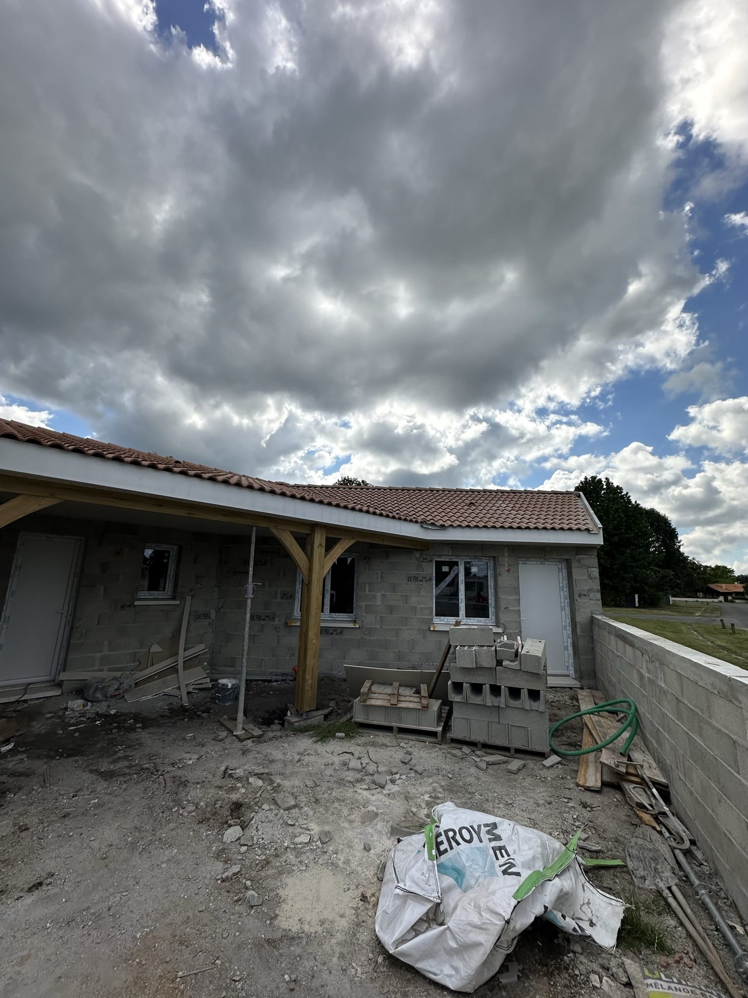 Chantier de maison en parpaings, toit de tuiles et sac Leroy Merlin sous un ciel chargé.