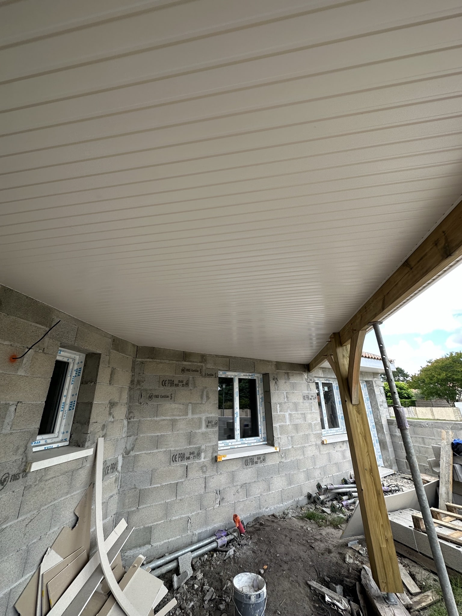 Mur en parpaings, fenêtres blanches et plafond en lambris PVC blanc sous auvent bois.